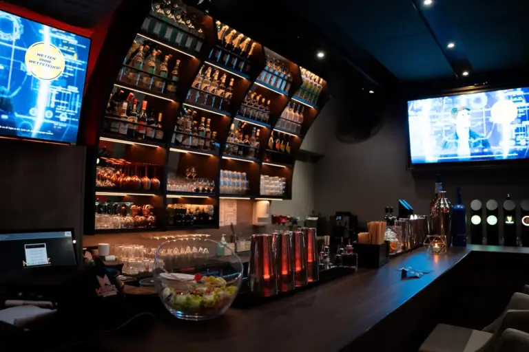 Eine schlichte, moderne Bar mit Regalen voller Spirituosenflaschen, Gläser und Cocktailshaker. Auf großen Bildschirmen werden Grafiken angezeigt, und auf dem Tresen steht eine Schale mit Früchten, die von einer stimmungsvollen Beleuchtung begleitet wird.