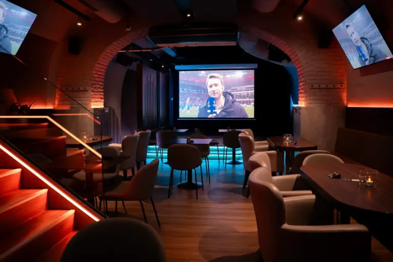 Eine schummrig beleuchtete Lounge mit modernen Stühlen und Tischen steht vor einem großen Bildschirm mit Sportübertragungen. Das Ambiente bietet warme Beleuchtung, freiliegende Backsteinbögen und eine gemütliche Atmosphäre.