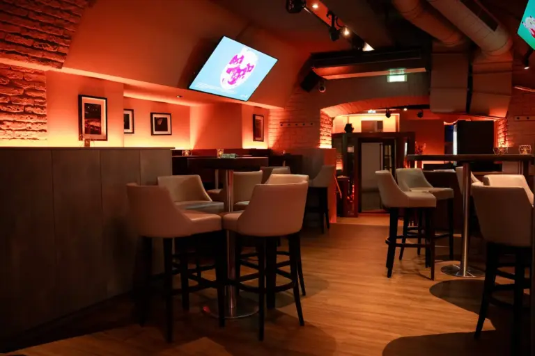 Eine moderne Bar mit warmer Beleuchtung, hohen Tischen und gepolsterten Stühlen, Holzböden, freiliegenden Ziegelwänden, gerahmten Fotos und einem großen, an der Decke montierten Fernseher mit einer bunten Grafik.