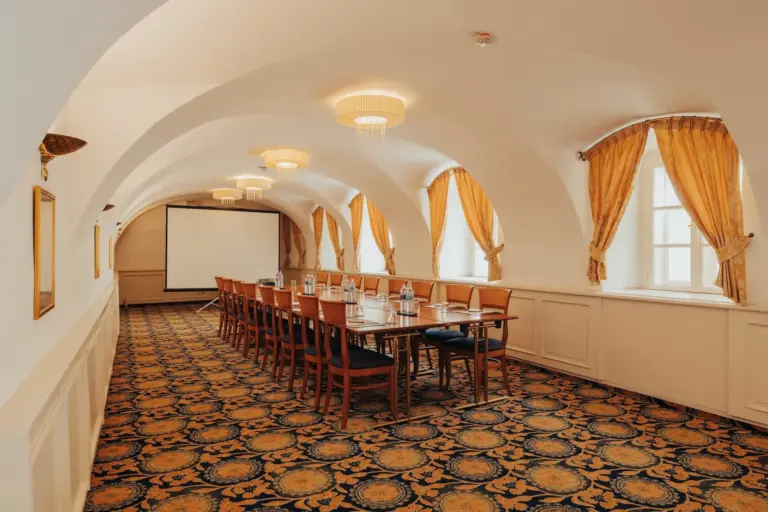 Ein langer Konferenztisch mit Stühlen steht in einem eleganten Raum mit gewölbten Decken, gemustertem Teppich, Kronleuchtern, goldenen Vorhängen und einer Projektionsfläche am anderen Ende.
