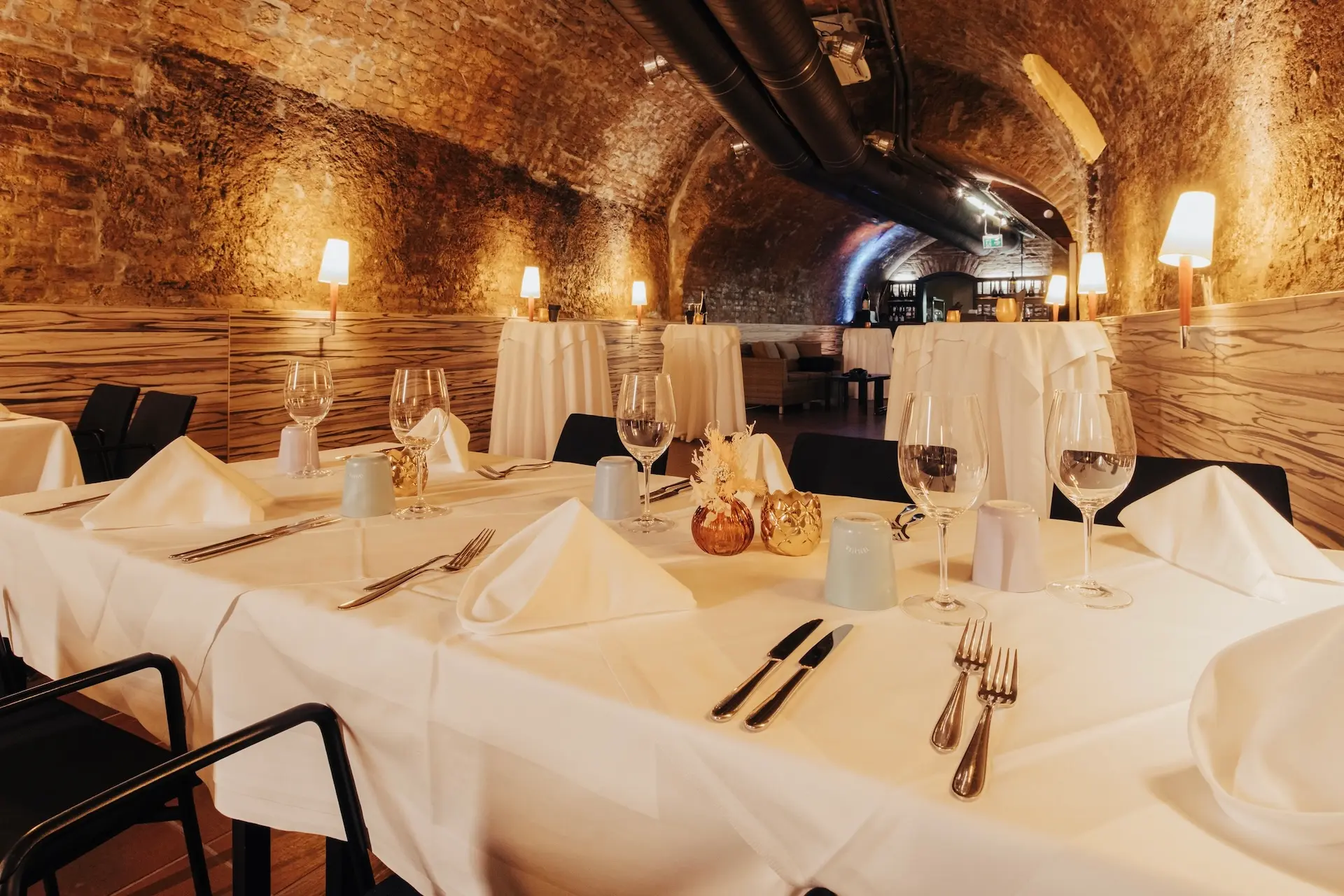 Elegantes Restaurantinterieur mit gewölbter Ziegelsteindecke, sanfter, warmer Beleuchtung und mit weißen Tischdecken, Servietten, Weingläsern und Besteck gedeckten Tischen, die eine einladende und anspruchsvolle Atmosphäre schaffen.