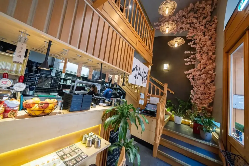 Ein modernes japanisches Restaurant mit Holzdekor, hängenden Laternen, Topfpflanzen und einem Kirschblütenbaum an der Wand in der Nähe einer Treppe, die zu einer oberen Etage führt. Die offene Küche ist auf der linken Seite zu sehen.