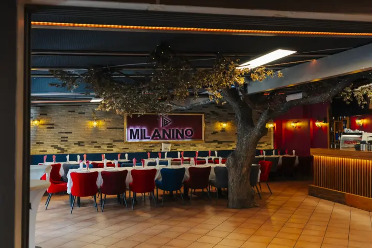 Hauptraum im Milanino in Wien Simmering mit großem Dekobaum, gedeckten Tafeln, roten und blauen Samtsesseln und beleuchtetem Milanino-Schriftzug an der Ziegelwand, rechts die Bar