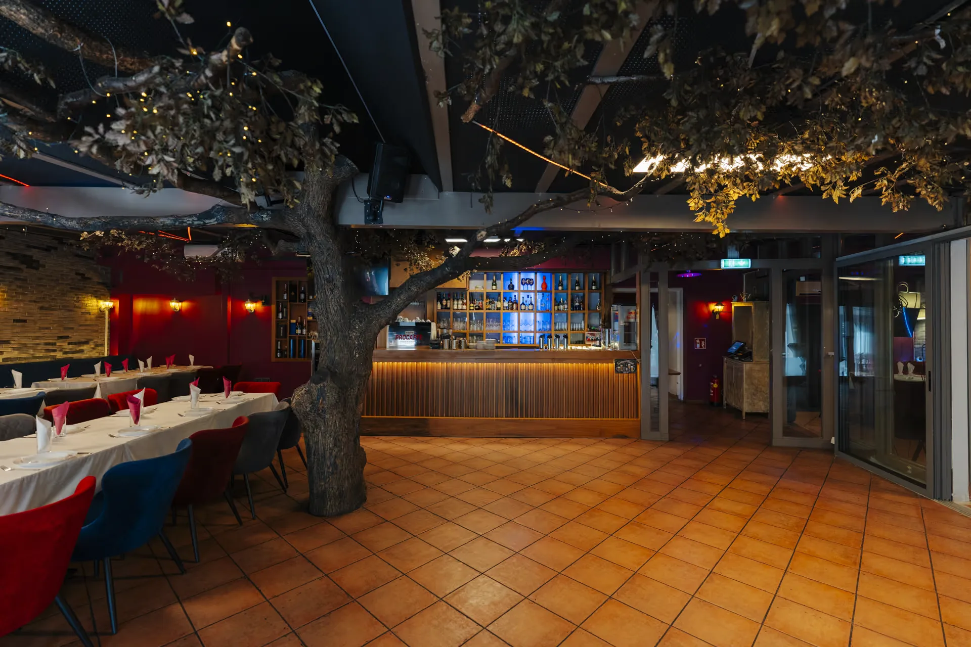Bar im Milanino mit Holzfront, warmer Beleuchtung und blau beleuchtetem Flaschenregal, daneben der Dekobaum und gedeckte Tische