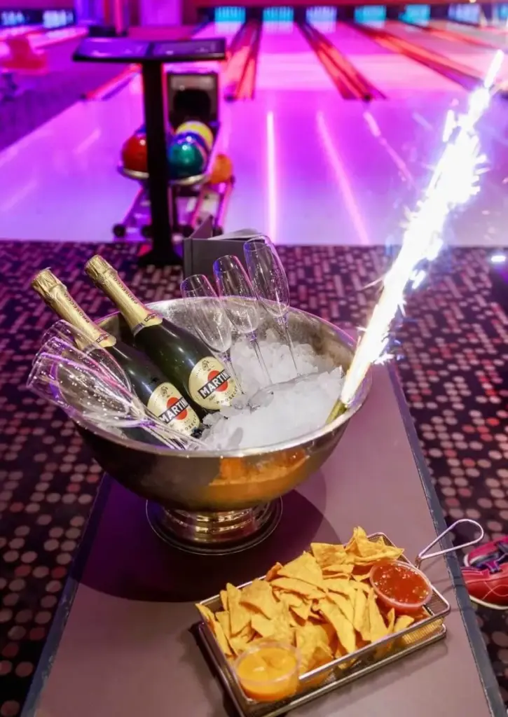 Eine Schale mit Champagnerflaschen, Gläsern und einer brennenden Wunderkerze steht auf einem Tisch neben Nachos und Dips im Millennium City Bowling, während im Hintergrund bunte Lichter und Bowlingbahnen leuchten.