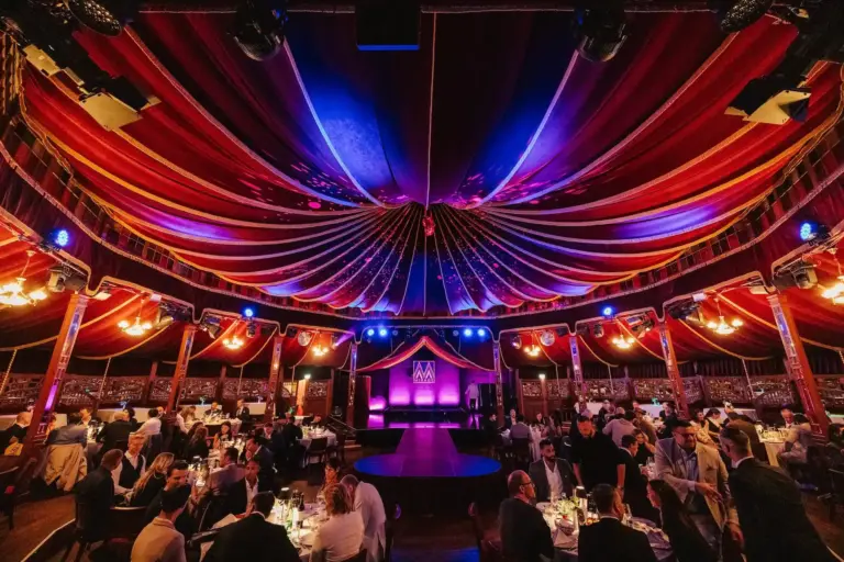 In einem großen, eleganten Zelt mit einer farbenfrohen, drapierten Decke sitzen die Gäste rund um eine runde Bühne, die von violetten, roten und blauen Lichtern beleuchtet wird.