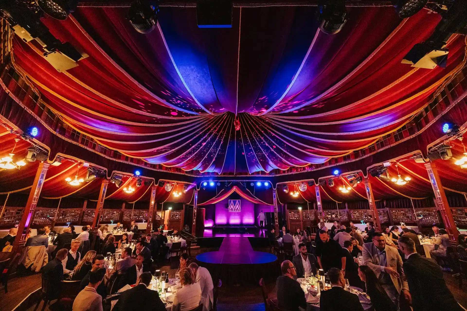In einem großen, eleganten Zelt mit einer farbenfrohen, drapierten Decke sitzen die Gäste rund um eine runde Bühne, die von violetten, roten und blauen Lichtern beleuchtet wird.