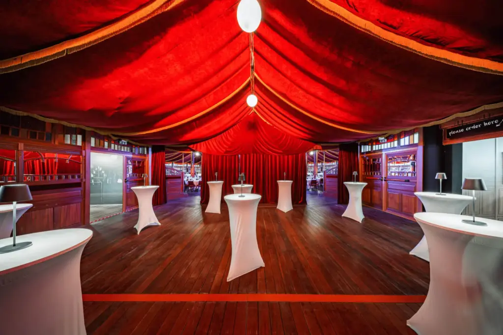 Ein großzügiger Veranstaltungsraum mit einer rot drapierten Decke, Holzböden und mit weißen Tüchern bedeckten Cocktailtischen unter Hängelampen, die eine elegante und festliche Atmosphäre schaffen.