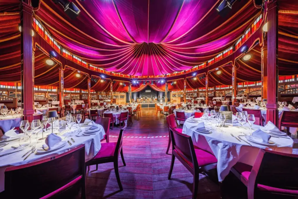 Mirage Ein üppiges Restaurant mit runden Tischen, eleganten roten Stühlen und einer dramatischen lila-roten Zeltdecke, die eine lebendige und luxuriöse Atmosphäre schafft.