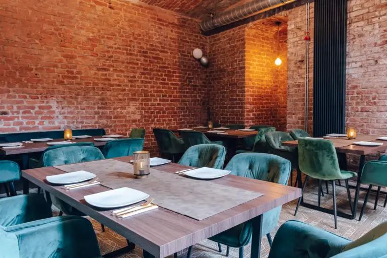 Ein gemütliches Restaurant mit freiliegenden Backsteinwänden, Holztischen mit weißen Tellern und Besteck, grünen Samtstühlen und warmer Beleuchtung.
