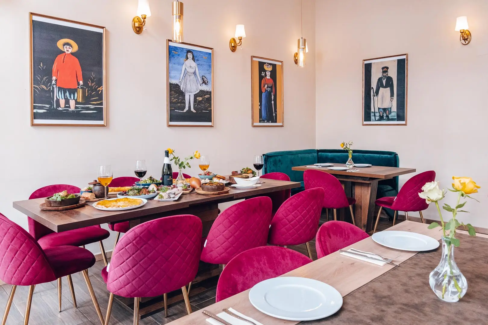Ein stilvoller Essbereich mit Tischen, die für eine Mahlzeit gedeckt sind, mit Stühlen in leuchtendem Magenta, grünen Sitznischen und gerahmten Porträts an den Wänden. Auf den Holztischen sind Teller mit Speisen und Getränken angerichtet.