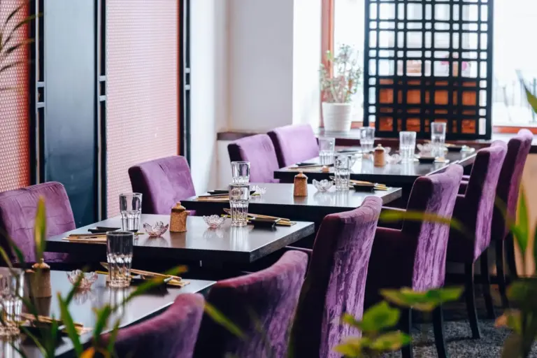 Ein modernes Restaurant mit gedeckten Tischen, Stühlen aus lila Samt, Gläsern und Besteck, neben großen Fenstern mit Tageslicht und einigen Zimmerpflanzen.
