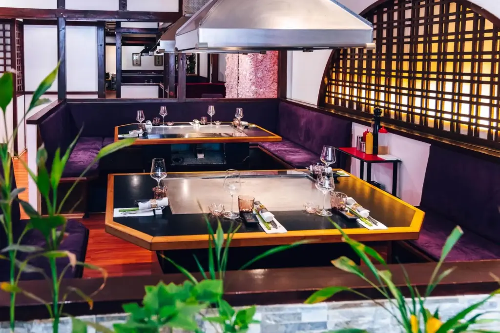 Ein modernes Restaurantinterieur mit zwei Teppanyaki-Tischen, die mit Weingläsern und Besteck gedeckt sind. Lila gepolsterte Sitzgelegenheiten umgeben die Tische. Grüne Pflanzen schmücken den Vordergrund, und warme Beleuchtung erfüllt den Raum.