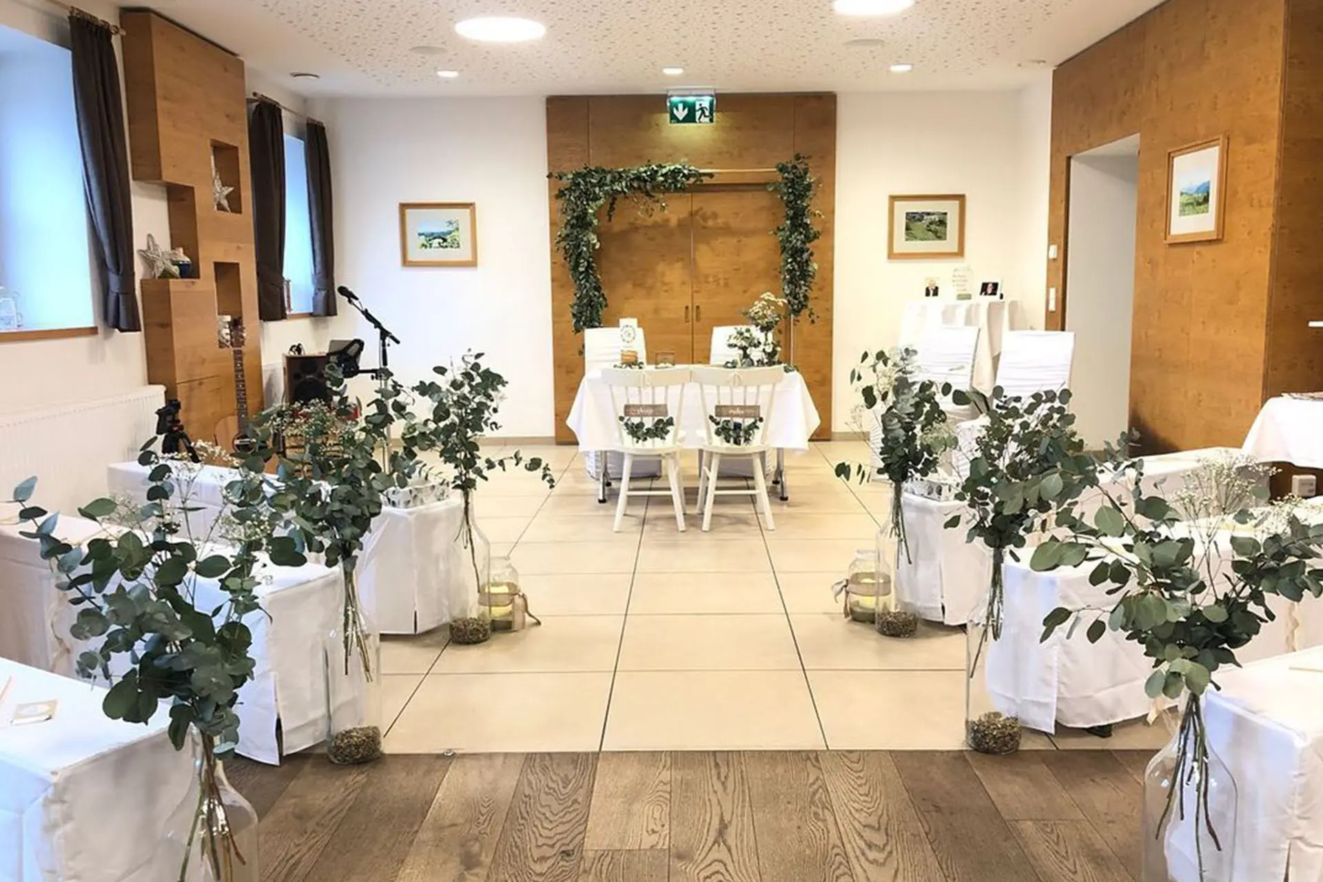 Eine kleine Indoor-Hochzeitszeremonie mit in zwei Reihen angeordneten Stühlen, dekoriert mit Grünzeug und weißen Decken. An der Vorderseite steht ein Tisch mit zwei Stühlen unter einem einfachen, mit grünem Laub geschmückten Bogen.