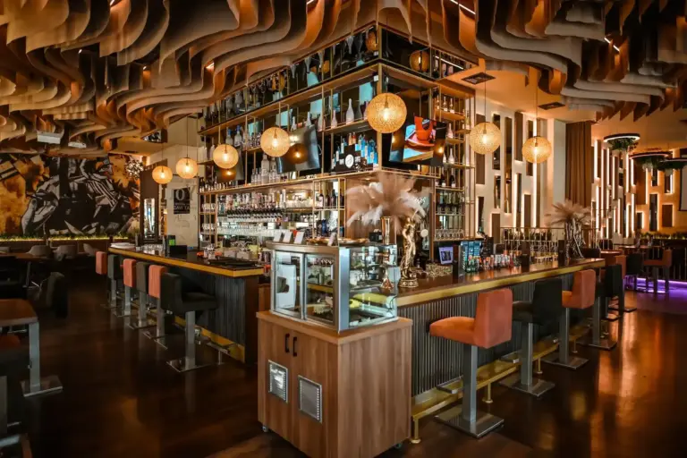 News Café Eine stilvolle Bar mit modernem Dekor, einer zentralen Inselbar, Regalen mit Flaschen, warmer Hängebeleuchtung, Barhockern aus Samt und gewellten Holzdecken, die eine gemütliche, gehobene Atmosphäre schaffen.
