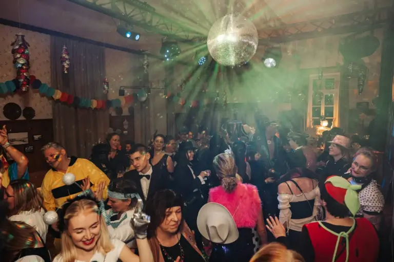 Eine lebhafte Menschenmenge in farbenfrohen Kostümen tanzt unter einer funkelnden Discokugel bei einer festlichen Indoor-Party, die mit Papiergirlanden und Lichtern geschmückt ist. Die Menschen lächeln und feiern in verschiedenen Outfits.
