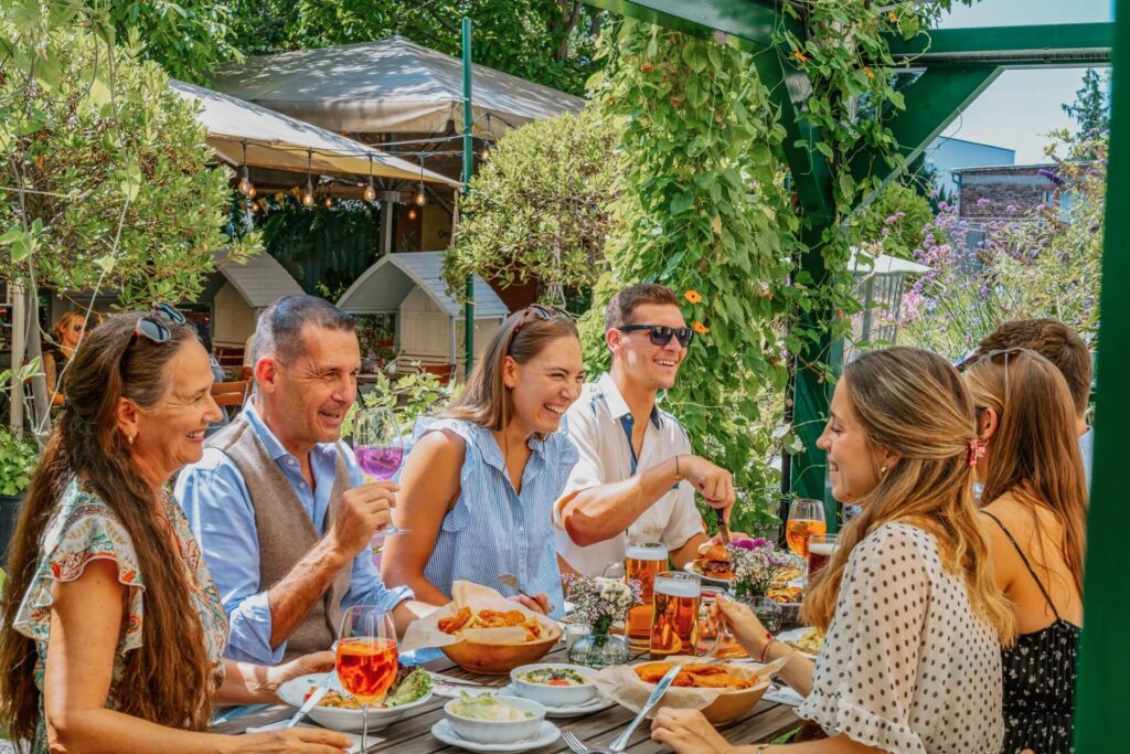 Eine Gruppe von Menschen sitzt an einem Tisch im Freien und genießt gemeinsam Speisen und Getränke. Sie lächeln und unterhalten sich, umgeben von Grün und Sonnenlicht, was eine lebhafte und fröhliche Atmosphäre schafft.