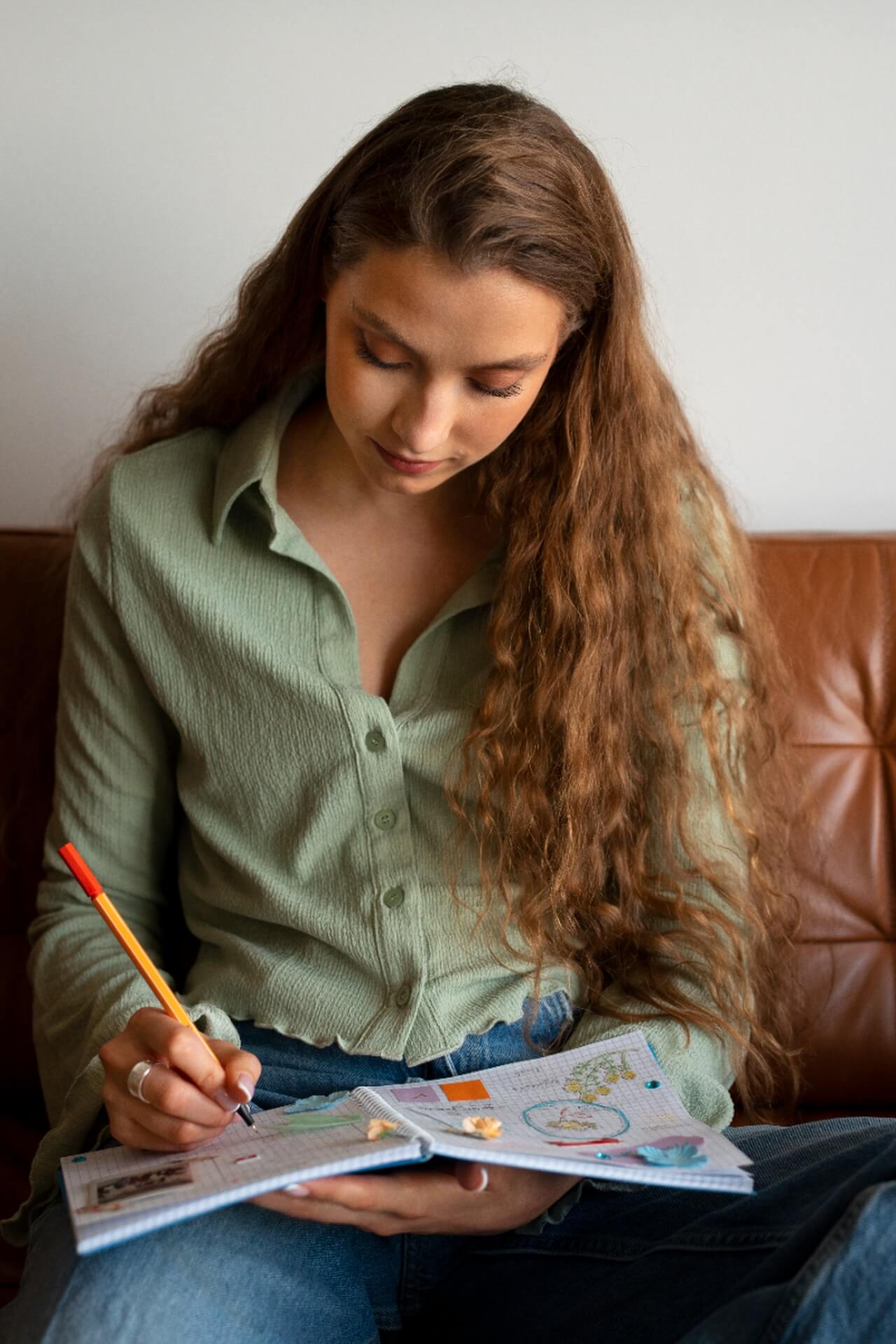 Eine junge Frau mit langen lockigen Haaren sitzt auf einer braunen Couch, schaut nach unten und schreibt mit einem gelben Stift Neujahrsvorsätze in ein Notizbuch. Sie trägt ein hellgrünes Button-up-Hemd und eine blaue Jeans.