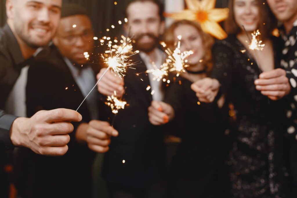 Eine Gruppe von Menschen in Partykleidung lächelt und hält Wunderkerzen in der Hand. Sie feiern die Neujahrsvorsätze in einer festlichen Umgebung mit einer großen gelben Blumendekoration im Hintergrund.