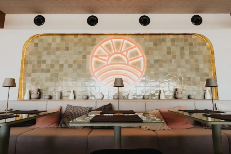 Modernes Restaurantinterieur mit gepolsterten Sitzbänken, dekorativen Kissen, minimalistischen Tischlampen und einer gekachelten Wand mit einem großen leuchtenden Neonsonnensymbol. Neutrale Farben und schlichte Töpferwaren schaffen eine gemütliche, stilvolle Atmosphäre.