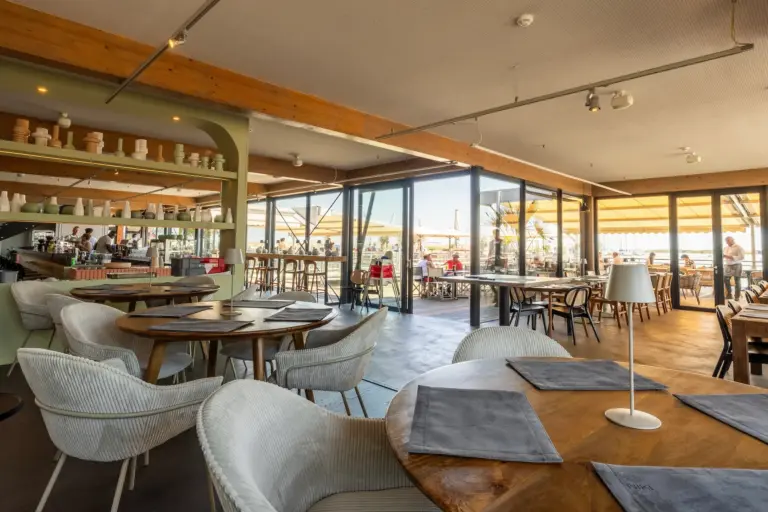 Ein modernes Restaurant mit Holztischen, gepolsterten Stühlen und großen Fenstern, die das Sonnenlicht hereinlassen. Die Gäste essen sowohl drinnen als auch draußen auf einer Terrasse mit Sonnenschirmen. Auf den Tischen stehen Gedecke mit Speisekarten.