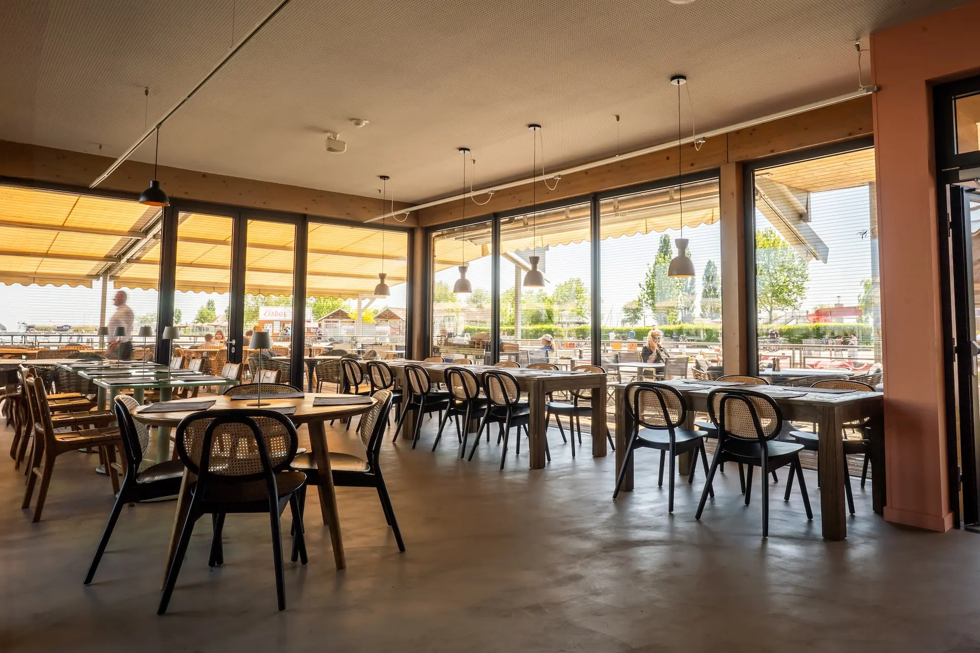 Ein geräumiges, modernes Restaurant mit großen Fenstern, die viel Tageslicht hereinlassen, ordentlich aufgestellten Holztischen und -stühlen und dem Blick auf eine Terrasse, auf der die Gäste draußen essen.