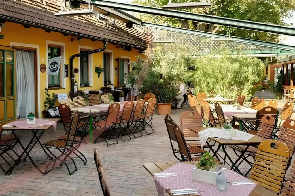 Die Terrasse des Restaurants ist mit Holztischen und -stühlen ausgestattet, die teilweise mit karierten Tischtüchern bedeckt sind. Das Gebäude ist gelb mit grünen Verzierungen, und Topfpflanzen schmücken den Bereich und schaffen eine gemütliche und einladende Atmosphäre.