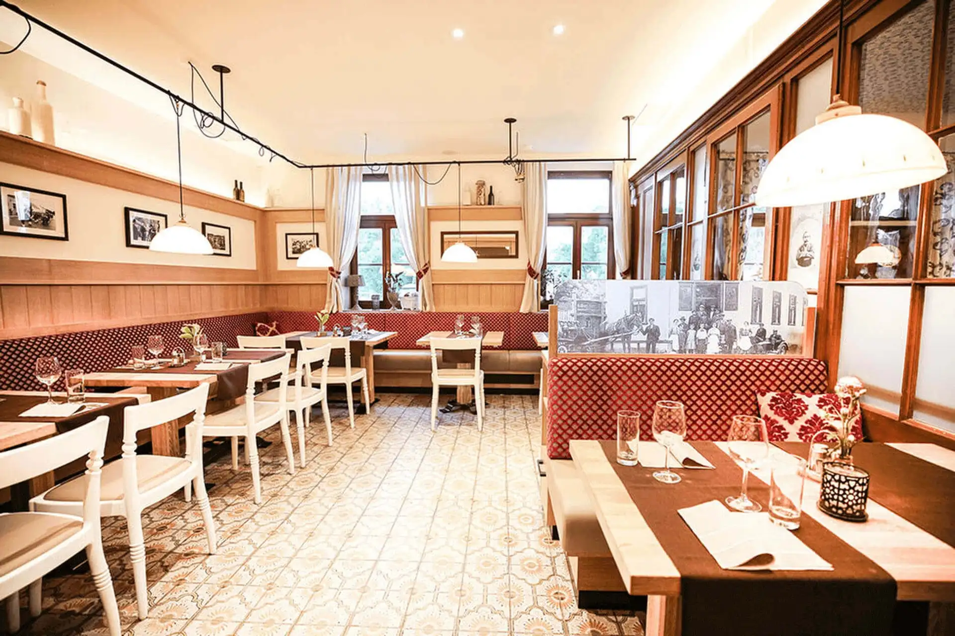 Ein helles, gemütliches Restaurant mit gemusterten Fliesenböden, roten gepolsterten Bänken, weißen Stühlen, Holztischen mit Gläsern und Servietten, Hängelampen und gerahmten Fotos an den Wänden.