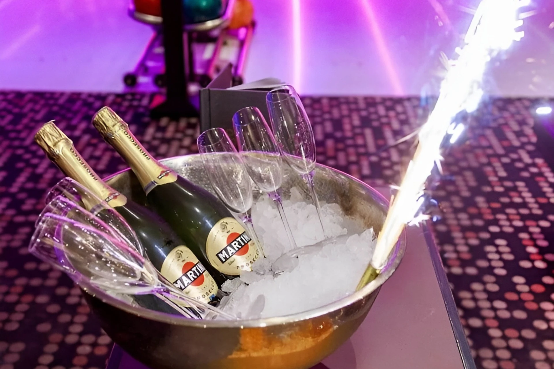 In einer mit Eis gefüllten Metallschüssel stehen drei Flaschen Martini-Sekt und mehrere leere Sektflöten. An einer Flasche ist eine brennende Wunderkerze befestigt, im Hintergrund leuchten Neonlichter und ein gemusterter Teppich.