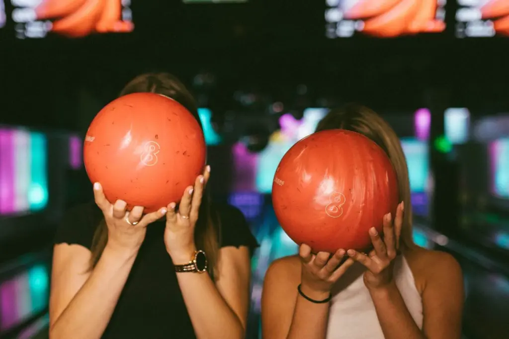 Zwei Personen stehen nebeneinander in einer Bowlingbahn und halten sich jeweils eine orangefarbene Bowlingkugel vor das Gesicht, wodurch ihre Gesichtszüge verdeckt werden. Der Hintergrund ist durch bunte Lichter unscharf.
