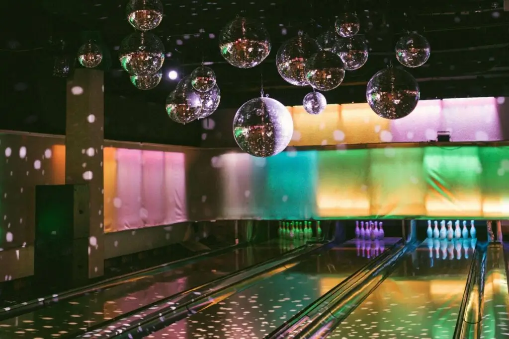 Eine Bowlingbahn mit glänzenden Bahnen, farbenfroher pastellfarbener Beleuchtung an den Wänden und mehreren von der Decke hängenden Discokugeln, die punktförmige Lichtmuster in den Raum werfen. Am Ende der Bahnen sind Bowlingkegel aufgestellt.