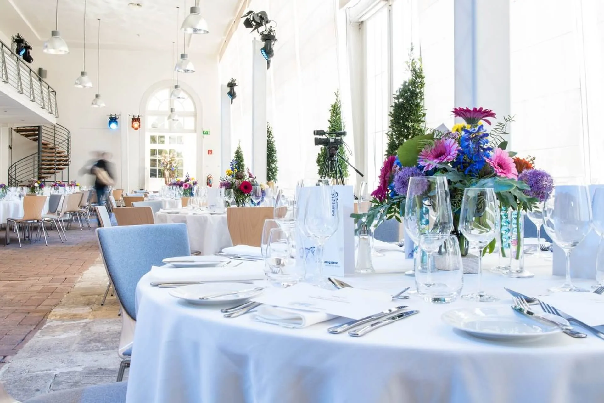 Ein heller, eleganter Bankettsaal für eine Veranstaltung mit runden Tischen mit weißen Tischdecken, Gläsern, Besteck und farbenfrohen Blumenkränzen. Große Fenster lassen natürliches Licht herein; im Hintergrund ist eine Treppe zu sehen.