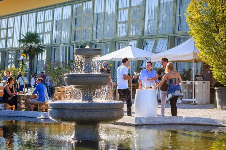 Bei einer Veranstaltung im Freien treffen sich die Menschen um einen Wasserbrunnen. Einige stehen an Tischen, während andere an einem sonnigen Tag in der Nähe eines Glasgebäudes und weißer Zelte sitzen und sich unterhalten. Bäume und Grünflächen umgeben die lebhafte Szene.