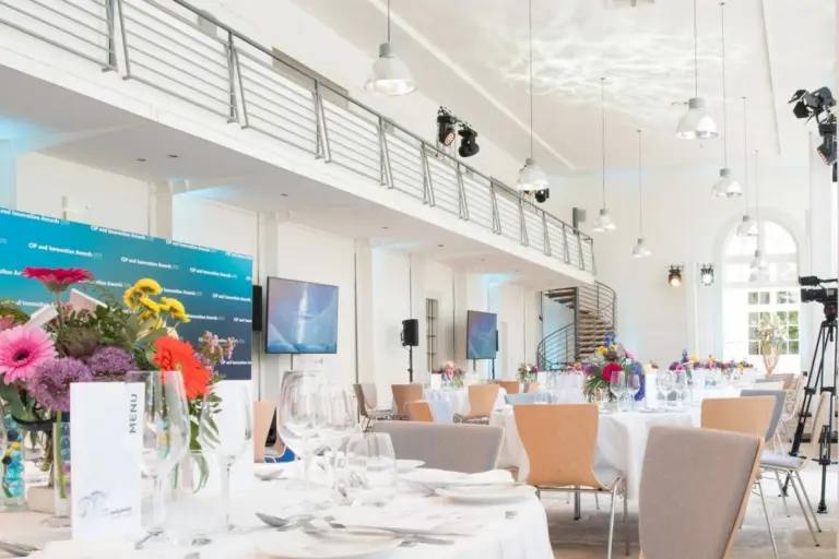 Ein heller, moderner Bankettsaal für eine Veranstaltung mit runden Tischen, weißen Tischdecken, Blumenschmuck, Gläsern und Speisekarten. Es gibt große Fenster, eine Wendeltreppe und Scheinwerfer über dem Kopf.