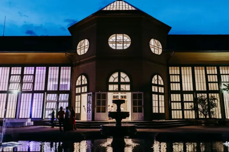 Ein großes, elegantes Gebäude mit hohen, beleuchteten Fenstern in der Abenddämmerung. Menschen stehen in der Nähe des Eingangs und eines zentralen Springbrunnens, der sich in einem Becken im Vordergrund spiegelt.