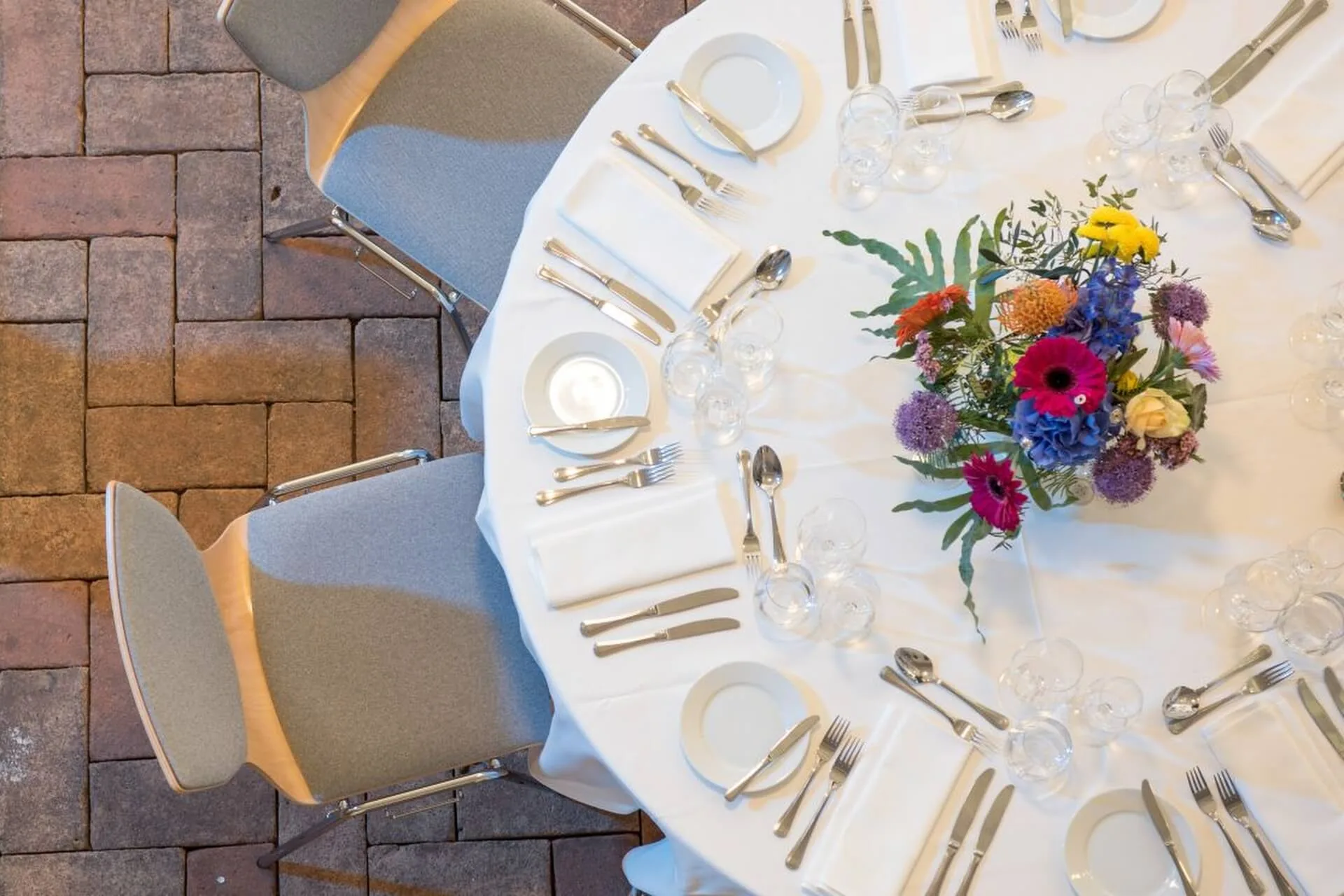 Draufsicht auf einen runden Tisch, der für eine Mahlzeit gedeckt ist, mit weißer Tischdecke, grauen Stühlen, Tellern, Besteck, Gläsern und einem bunten Blumenschmuck. Der Tisch steht auf einem Backsteinboden.