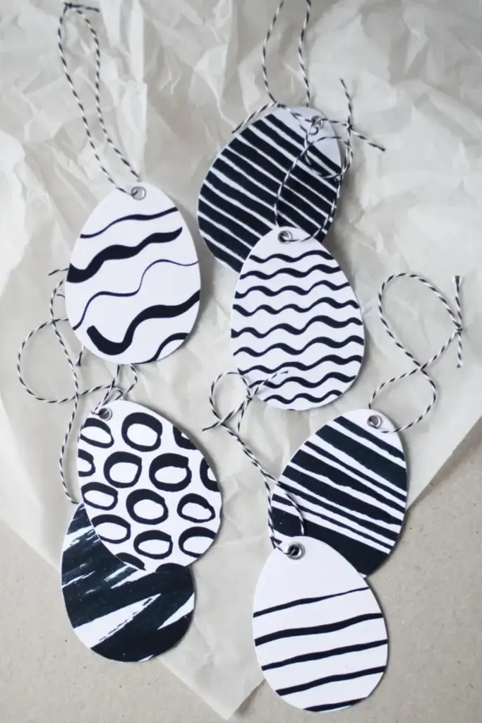 Sechs ovale Anhänger mit abstrakten Mustern in Schwarz und Weiß, darunter Wellenlinien, Kreise und Streifen, sind auf zerknittertem Papier angeordnet. Perfekt für Osterdeko-Basteln. Jeder Anhänger hat eine passende schwarz-weiße Schnurschlaufe zum Aufhängen.