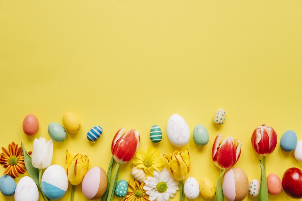 Bunte Ostereier färben und Frühlingsblumen, einschließlich Tulpen und Gänseblümchen, angeordnet entlang der Unterseite eines leuchtend gelben Hintergrunds mit Platz darüber für Text.