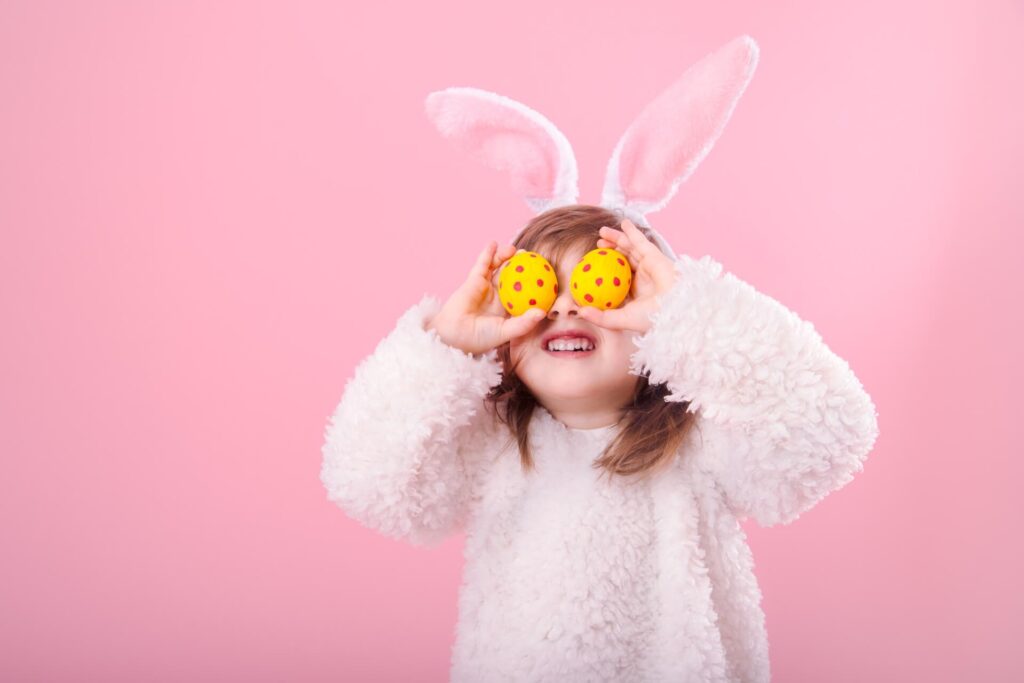 Ein Kind, das ein flauschiges weißes Kostüm und rosa Hasenohren trägt, hält zwei gelbe gepunktete Ostereier über die Augen und lächelt vor einem verspielten rosa Hintergrund.