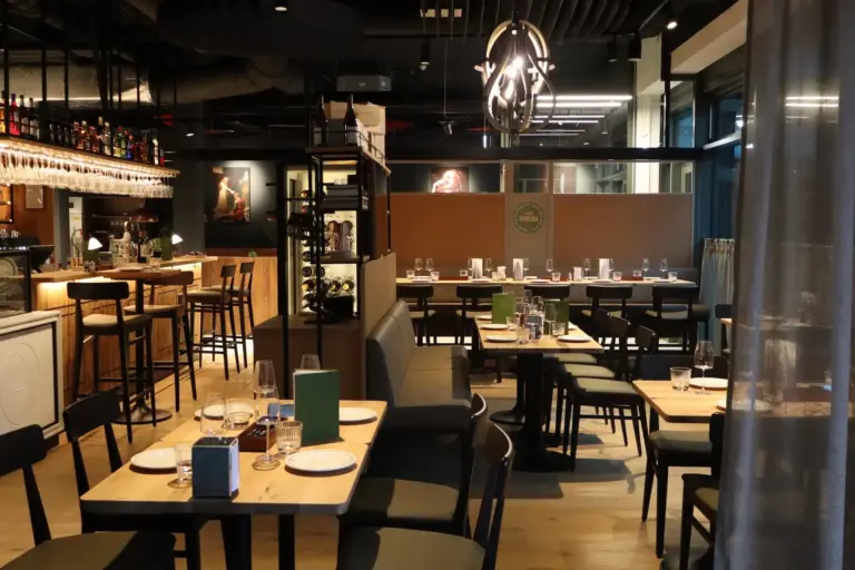 Ein modernes, stilvolles Restaurantinterieur mit Holztischen zum Essen, schwarzen Stühlen, einem Barbereich auf der linken Seite, gedämpfter Beleuchtung und zeitgenössischem Dekor. Der Raum scheint bereit zu sein, Gäste zu empfangen.