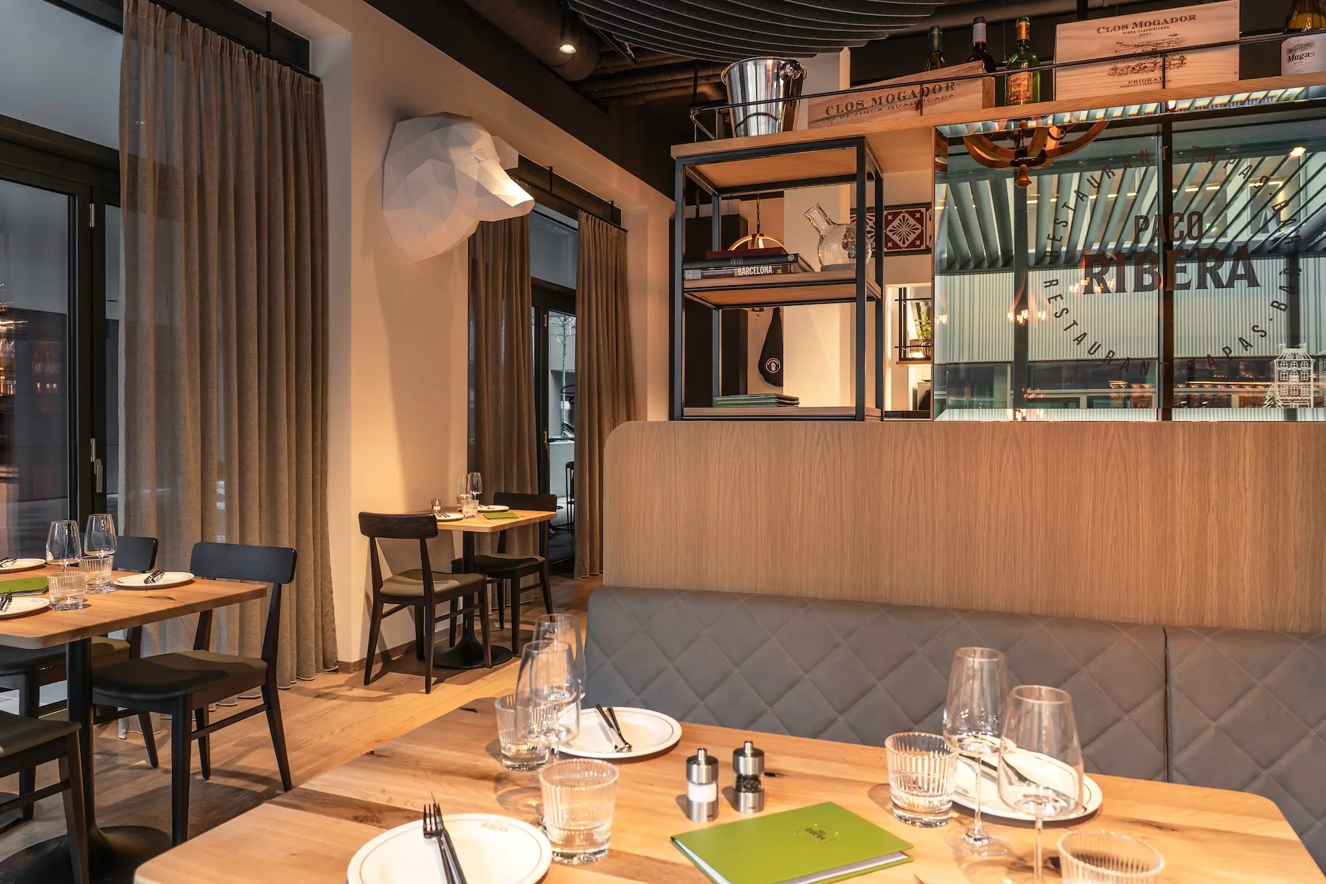 Modernes Restaurantinterieur mit Holztischen zum Essen, gepolsterten Sitznischen, Gläsern und Tellern. Ein Regal mit Flaschen und Kisten trennt den Essbereich ab, und große Fenster lassen natürliches Licht herein.