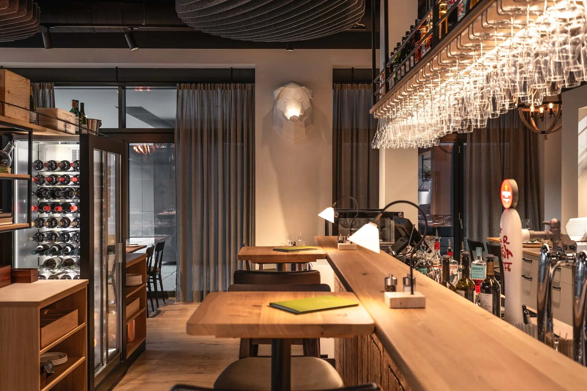 Eine gemütliche moderne Bar mit Holztresen, hohen Hockern, hängenden Weingläsern, einem Weinkühlschrank und warmer Beleuchtung. Regale mit Flaschen und eine Bierzapfanlage sind zu sehen, und große Fenster mit Vorhängen lassen natürliches Licht herein.