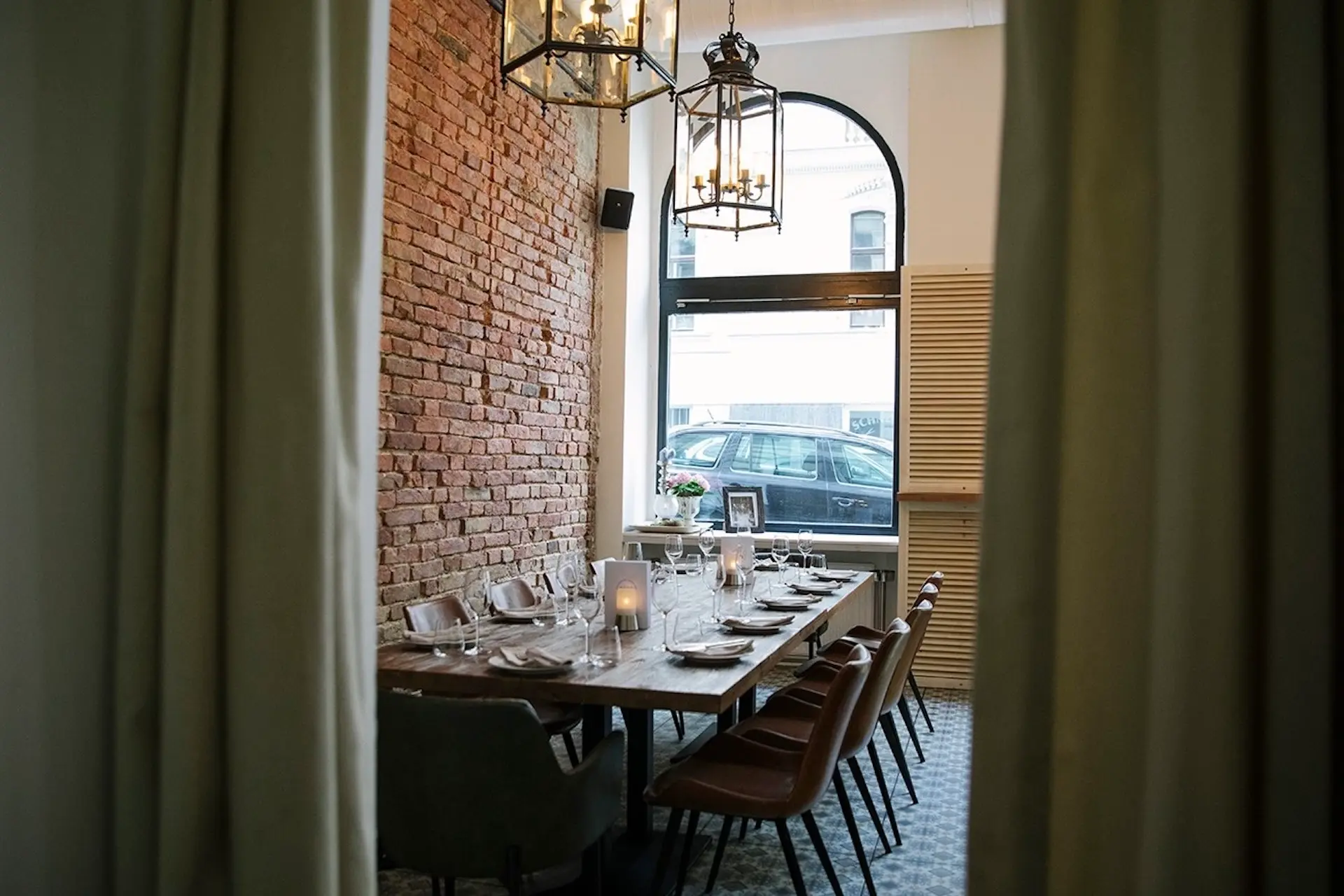 Ein gemütliches Esszimmer mit einem langen Holztisch von Paco, an dem eine Mahlzeit eingenommen wird, umgeben von braunen Stühlen. Die freiliegende Ziegelwand, das große Fenster, der gemusterte Fußboden, die Hängeleuchten und eine brennende Kerze schaffen eine warme, einladende Atmosphäre.