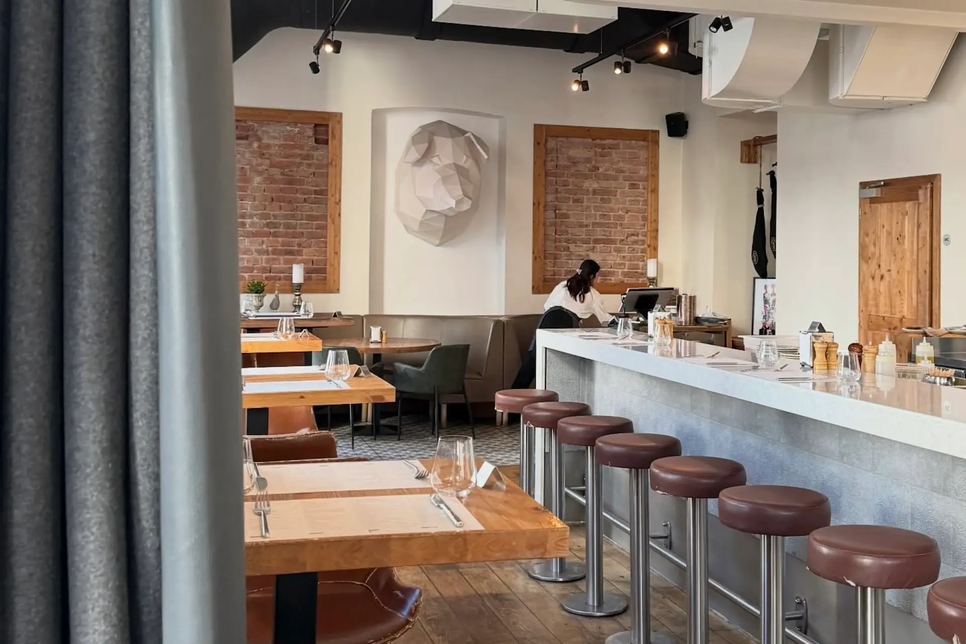 Ein modernes Restaurant mit Holztischen, braunen gepolsterten Barhockern am Tresen, freiliegenden Ziegelwänden und minimalistischem Dekor. Hinter dem Tresen arbeitet Paco in dem gut beleuchteten Raum.