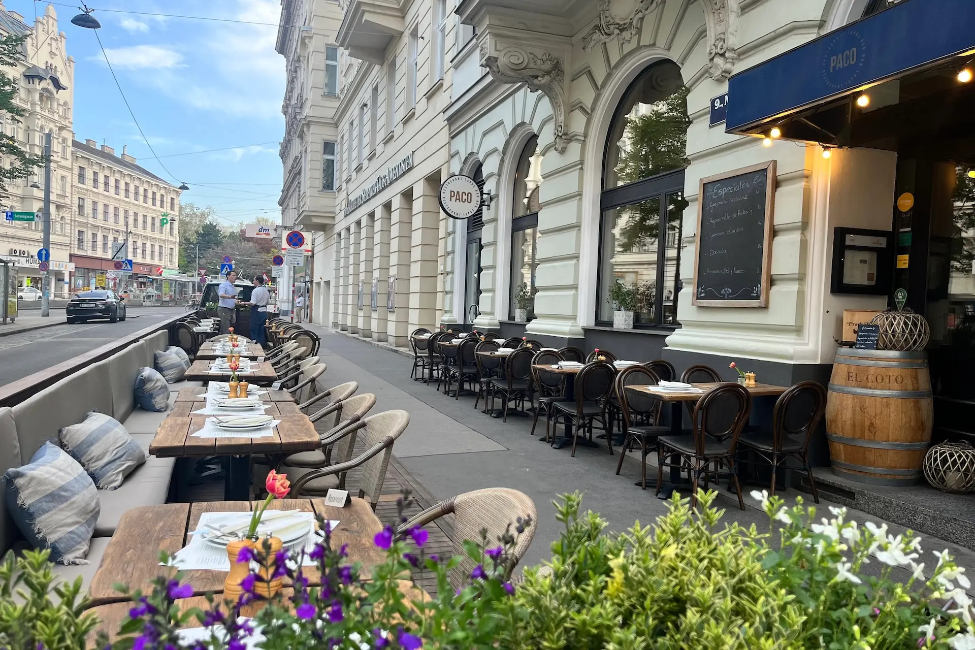 Sitzgelegenheiten im Freien in einem europäischen Café mit Holztischen und schwarzen Stühlen auf einem Bürgersteig in der Stadt, bunte Blumen im Vordergrund und historische weiße Gebäude entlang der Straße.