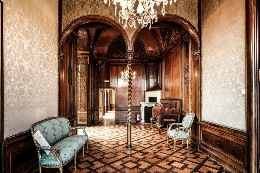 Der elegante Raum mit seiner kunstvollen Holzvertäfelung, dem gemusterten Parkettboden und den blauen Vintage-Polstermöbeln erinnert an die Pracht des Palais Auersperg. Eine gewölbte Tür rahmt einen Kristalllüster ein, während ein kleiner Kamin und ein antikes Klavier den Hintergrund zieren.