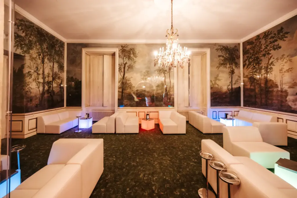 Eine elegante Lounge mit weißen Sesseln und beleuchteten Tischen, ein grüner Teppich, ein kunstvoller Kronleuchter und malerische Wandgemälde, die Bäume und die Natur darstellen, vermitteln das mondäne Ambiente des Palais Auersperg und schaffen eine einladende Atmosphäre.