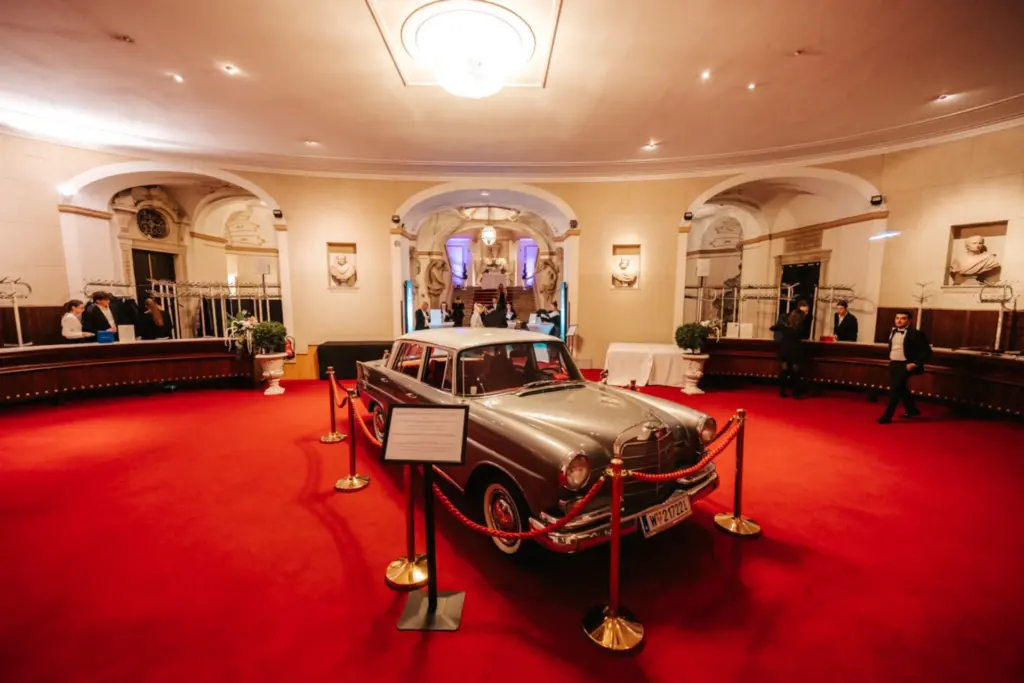 Ein silberner Oldtimer wird auf einem roten Teppich in den eleganten, hohen Sälen des Palais Auersperg ausgestellt, mit Kronleuchtern, dekorativen Bögen und Menschen, die um Samtbänder und ein Schild herumstehen, das den Wagen umgibt.