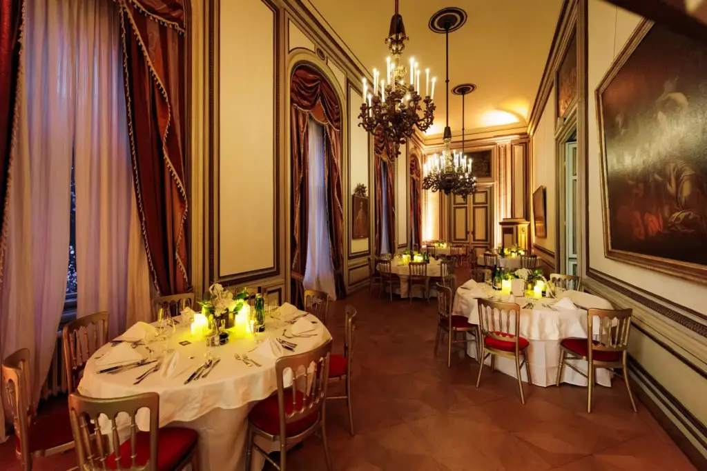 Eleganter Speisesaal mit runden Tischen, die für eine formelle Veranstaltung im Palais Auersperg gedeckt sind, mit weißen Tischtüchern, floralen Tafelaufsätzen und leuchtenden Kerzen. Große Fenster mit drapierten Vorhängen und verzierte Kronleuchter vervollständigen die luxuriöse Atmosphäre.