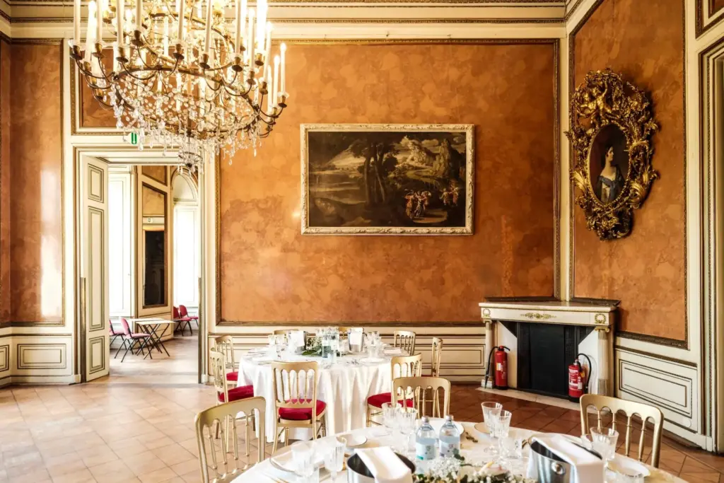 Ein eleganter Speisesaal im Palais Auersperg mit verzierten Wänden, Kronleuchtern, goldgerahmten Gemälden, einem Kamin und runden Tischen mit weißen Tischtüchern und Gläsern. Licht fällt durch große Türöffnungen im Hintergrund ein.
